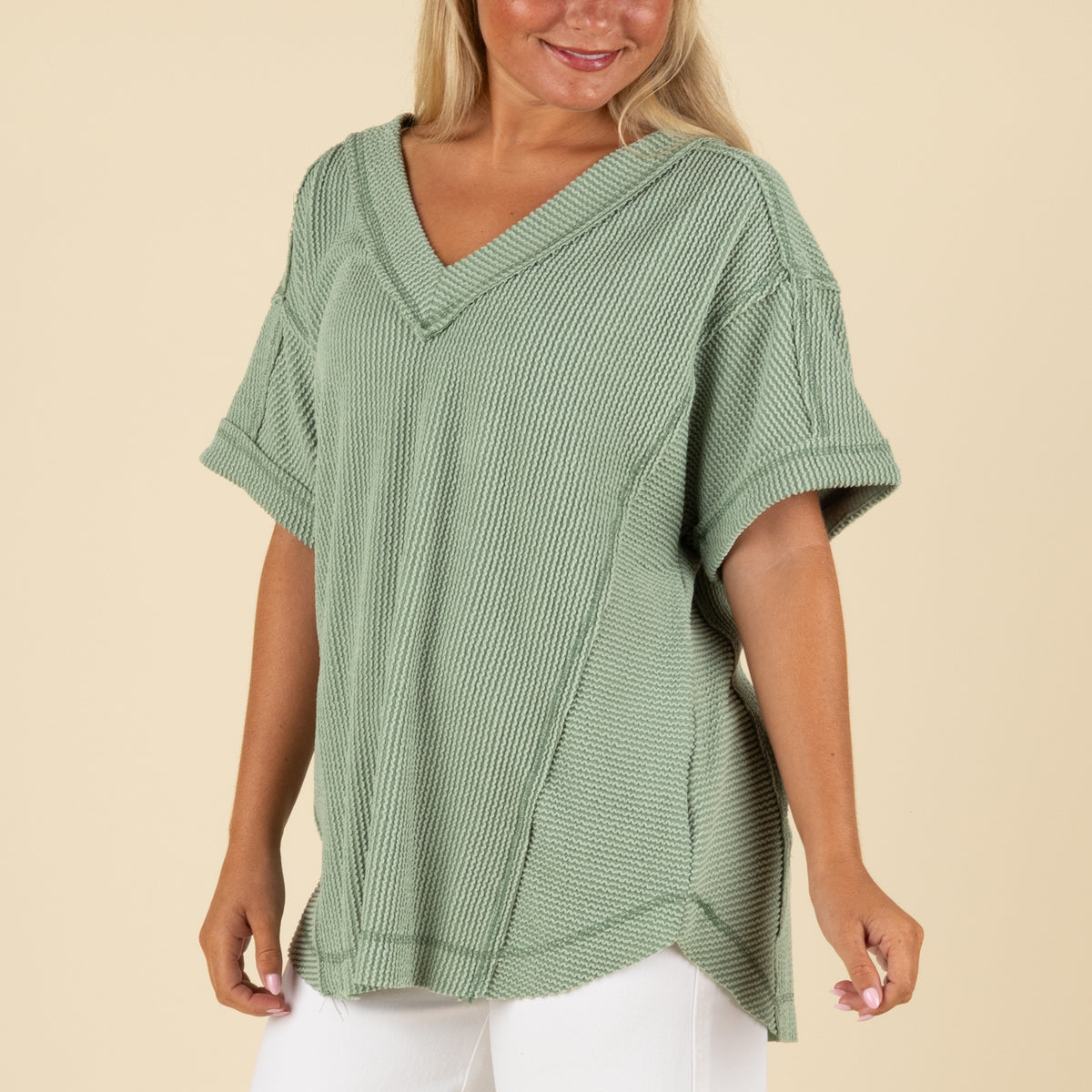 closeup front view of a woman wearing an oversized sage ribbed vneck top paired with white denim jeans.