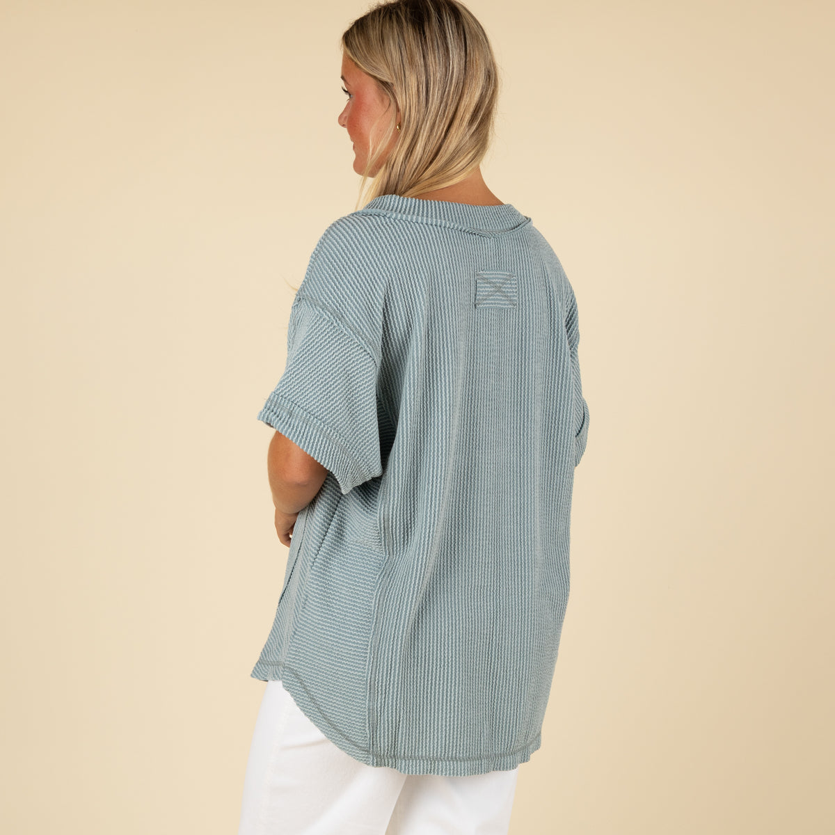 back view of a woman wearing an oversized slate blue ribbed vneck top paired with white denim jeans.
