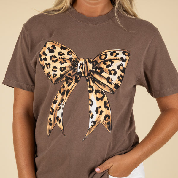 Brown t-shirt with a large leopard print bow design worn by a person.