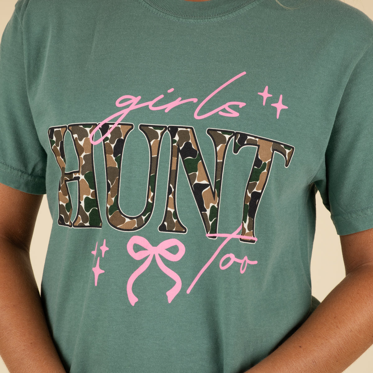 Green t-shirt with 'girls hunt too' text in pink and camouflage on a plain background