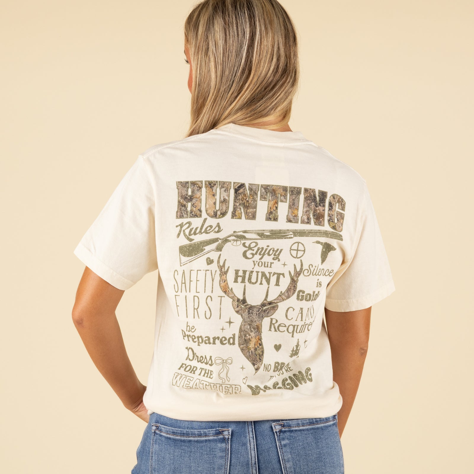 Back view of ivory t-shirt featuring camo deer graphic and large text reading “Hunting Rules” with various outdoor-themed phrases.