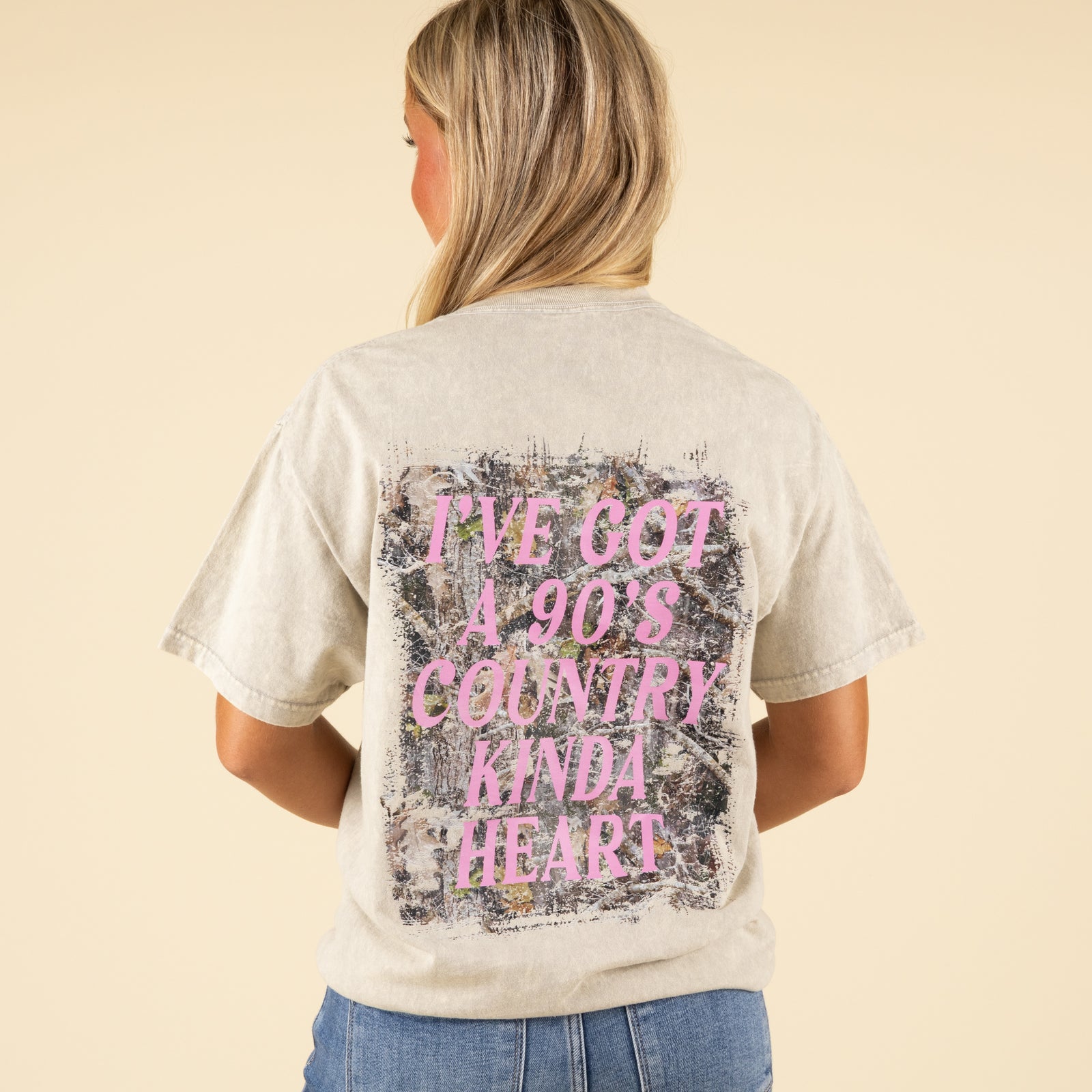 Back view of stonewashed beige t-shirt with distressed camo-print background and pink text reading “I’ve Got a 90’s Country Kinda Heart.”