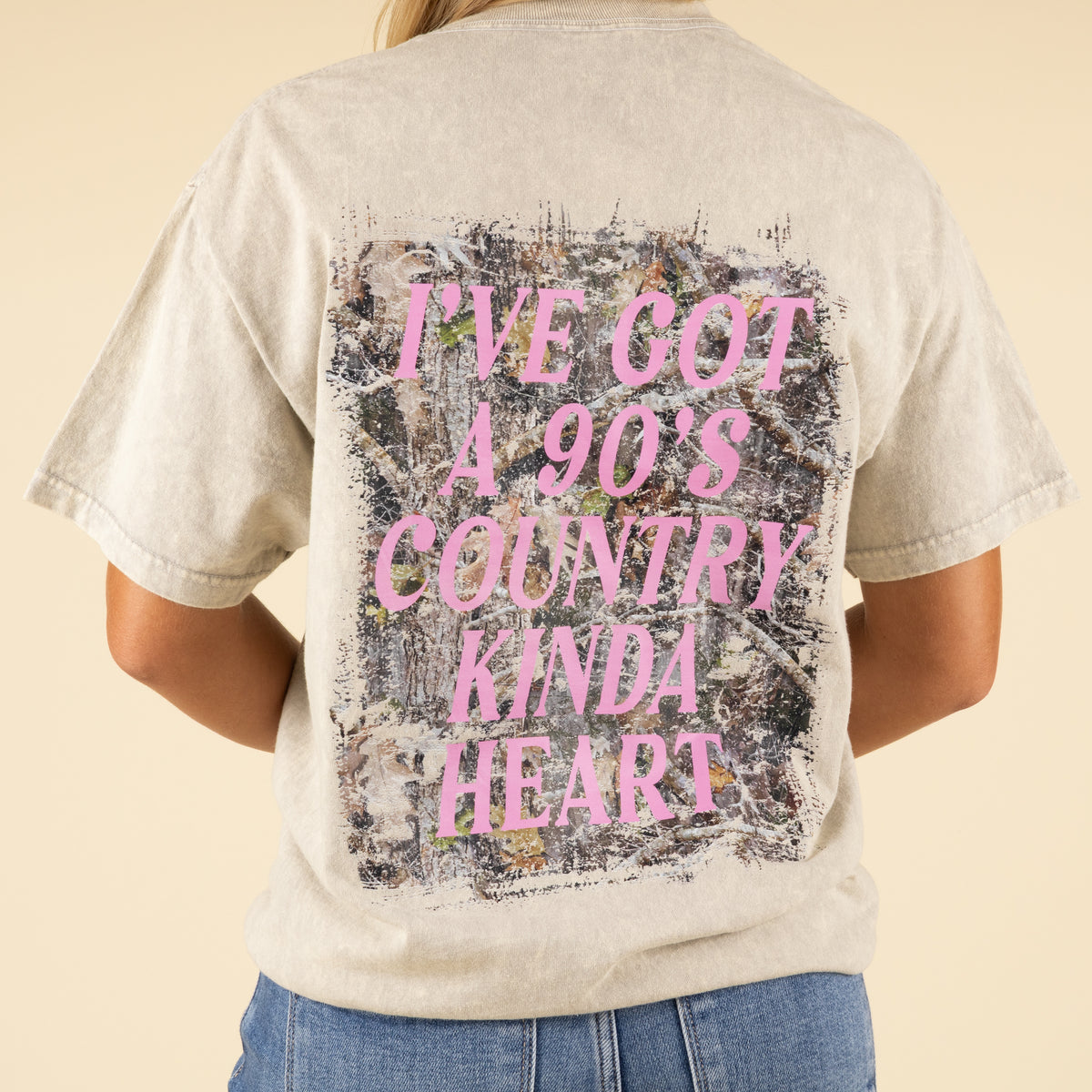 Close-up of back graphic showing camo background and bold pink “I’ve Got a 90’s Country Kinda Heart” text on beige t-shirt.