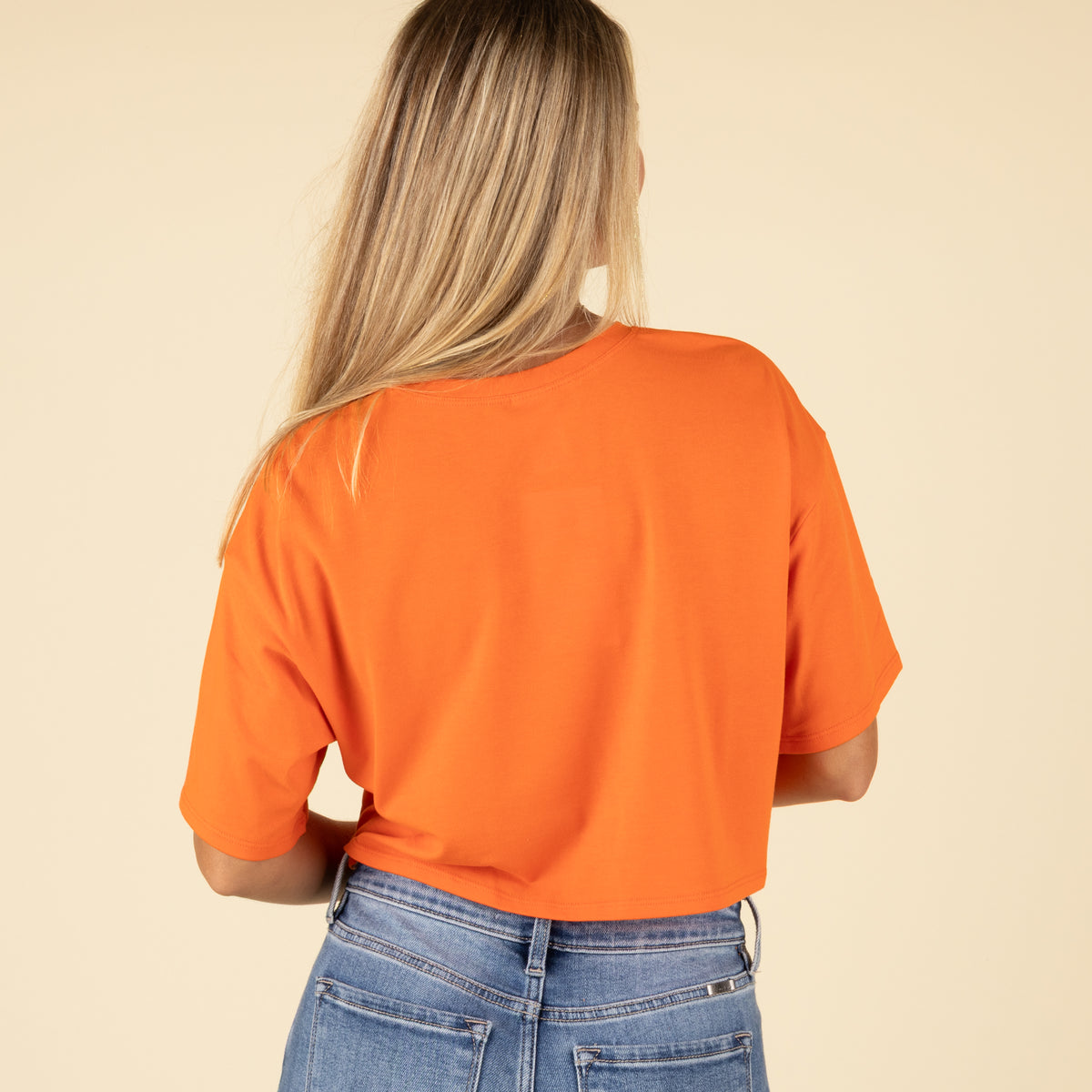 Back view of a woman wearing a solid orange cropped t-shirt with short sleeves and light-wash denim jeans.