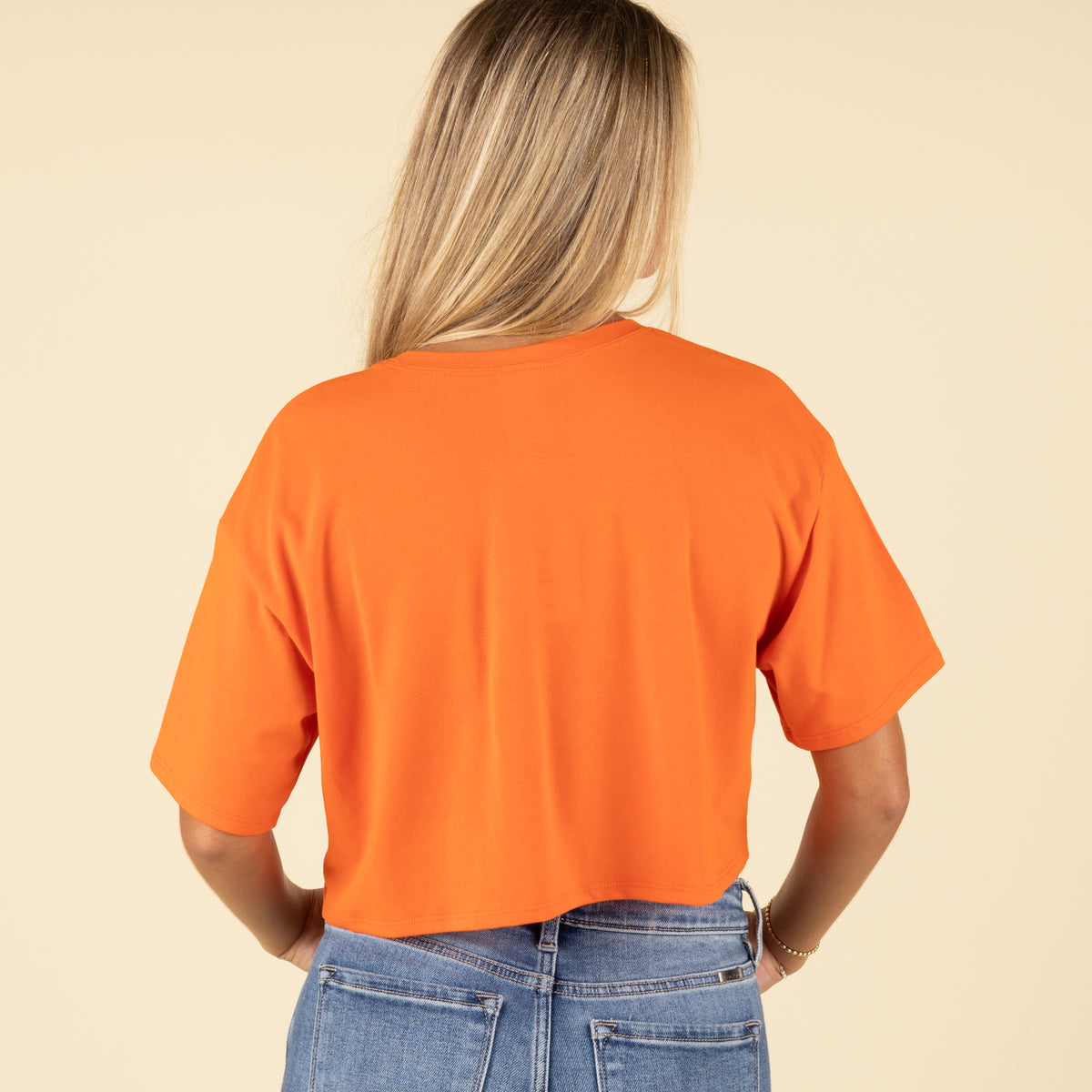 Back view of a woman wearing a solid orange cropped t-shirt with short sleeves and light-wash denim jeans.