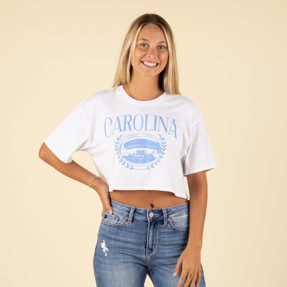 Front view of a woman wearing a white Carolina cropped t-shirt with a light blue collegiate graphic and light distressed denim jeans.