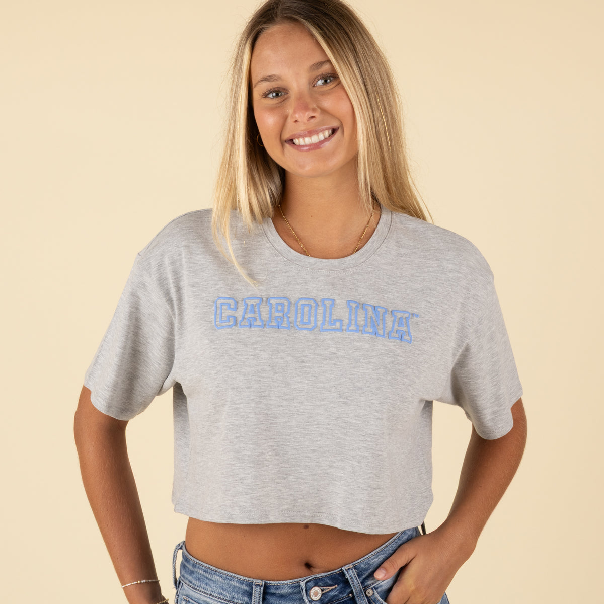 Close-up front view of a woman in a grey Carolina cropped t-shirt with light blue block lettering and short sleeves.