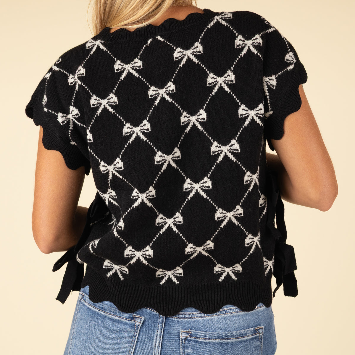 Back view of black knit sweater with white bow pattern and scalloped hem, styled with light wash jeans.