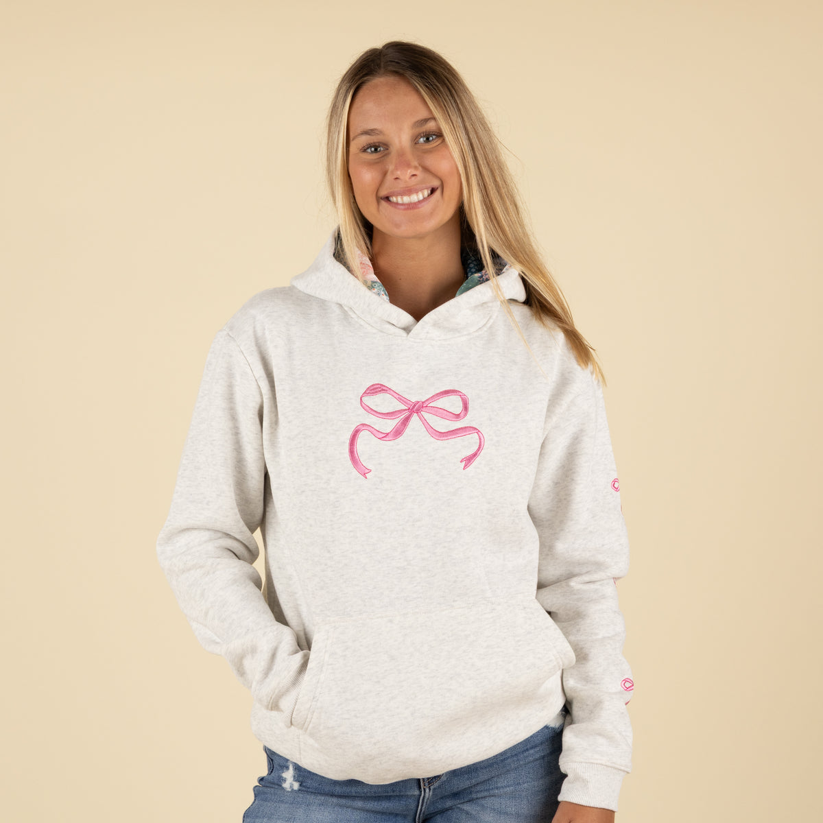 Front view of a woman wearing a light heather gray hoodie with a large pink bow embroidered on the chest.