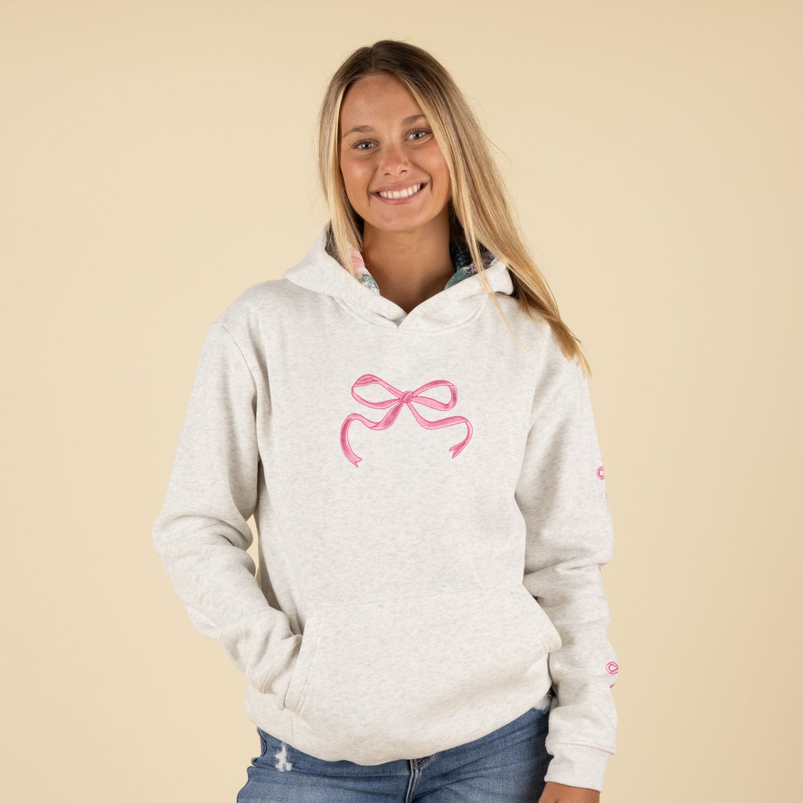 Front view of a woman wearing a light heather gray hoodie with a large pink bow embroidered on the chest.