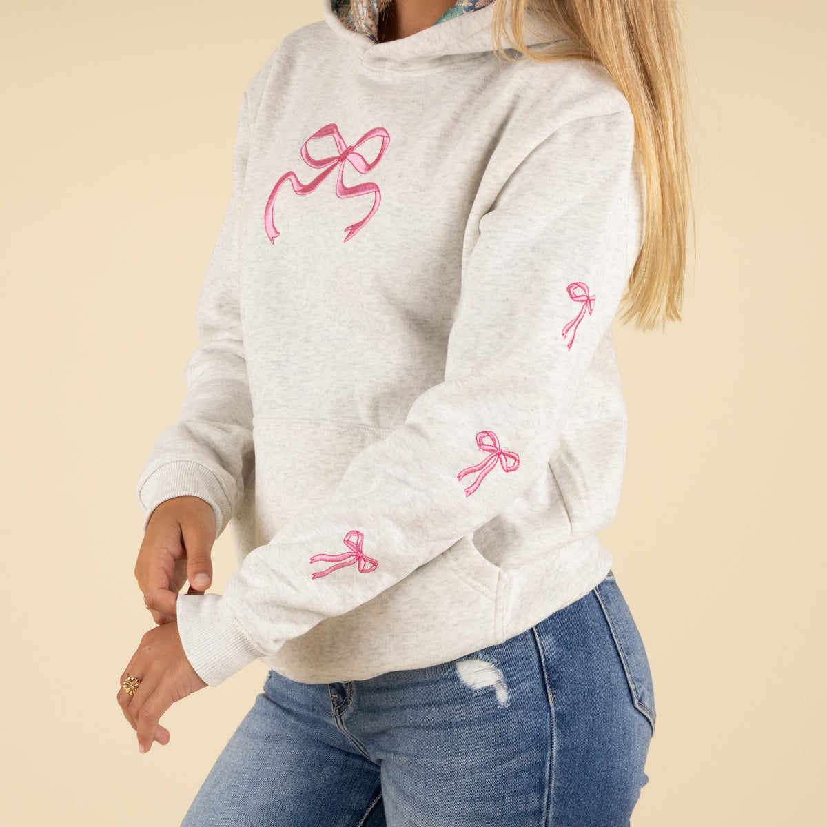 Side view of a woman wearing a light heather gray hoodie with multiple embroidered pink bows running along the sleeve.