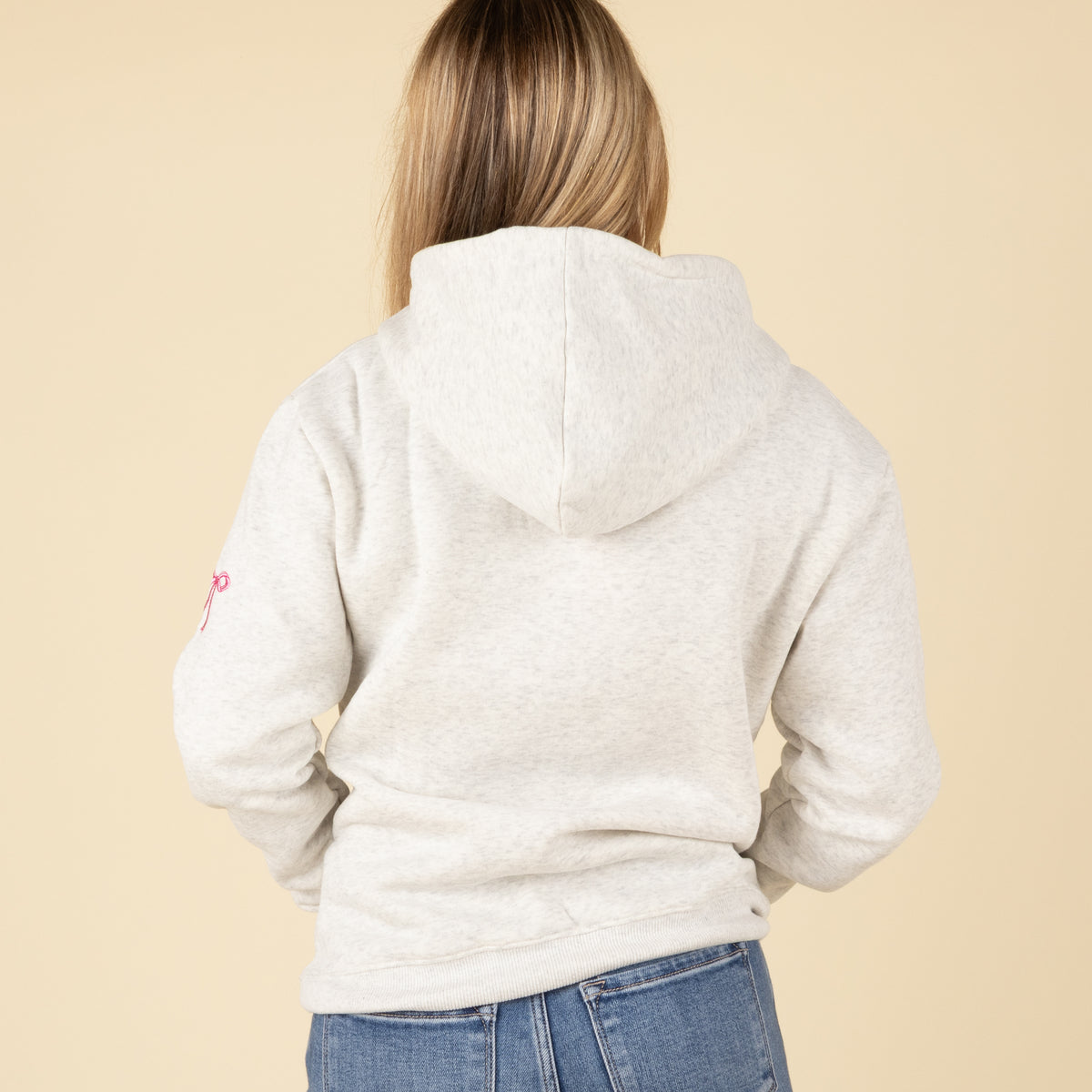 Back view of a woman wearing a light heather gray hoodie with the hood up, showing plain back and long sleeves with subtle bow detail on one arm.