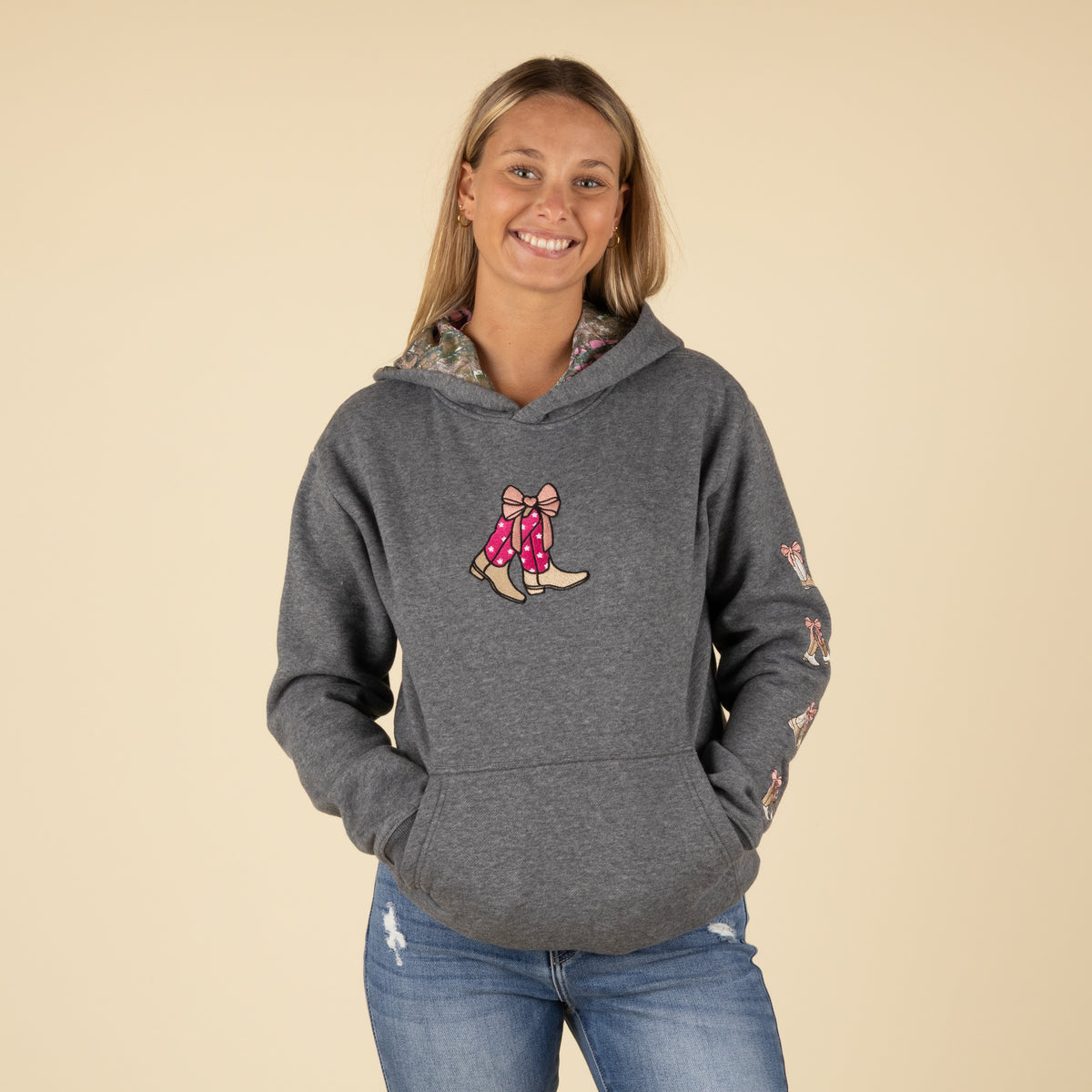 Front view of a woman wearing a dark gray hoodie with embroidered pink cowboy boots tied with a bow on the chest and boot embroidery on the sleeve.