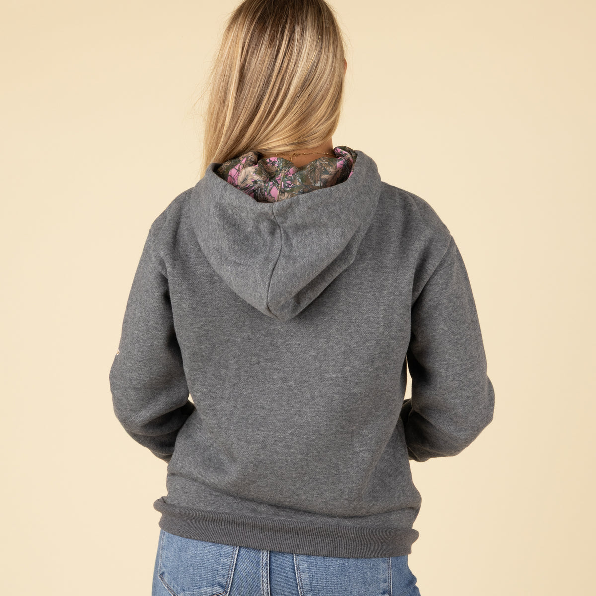 Full back view of the woman wearing the dark gray hoodie with a plain back and hood down, revealing the camo and pink lining inside.