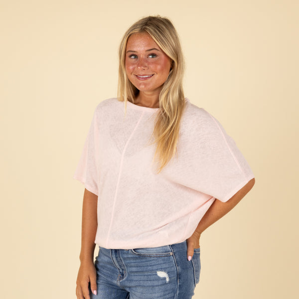  Woman wearing a light pink loose fit short sleeve top, angled side view with one hand in pocket.