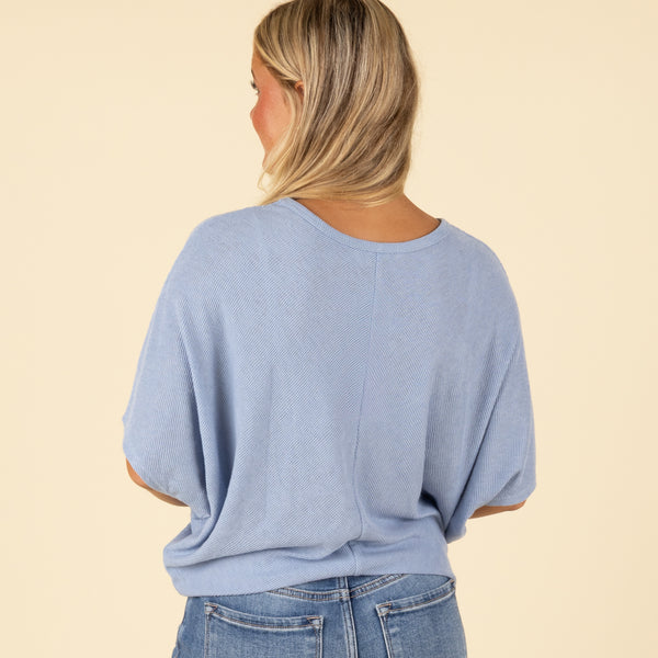  Back view of woman wearing a light blue loose fit short sleeve top with relaxed drape.