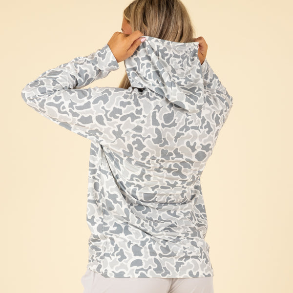 Back view of woman wearing grey and white camouflage hooded long sleeve shirt with light grey athletic shorts, lifting hood with both hands.