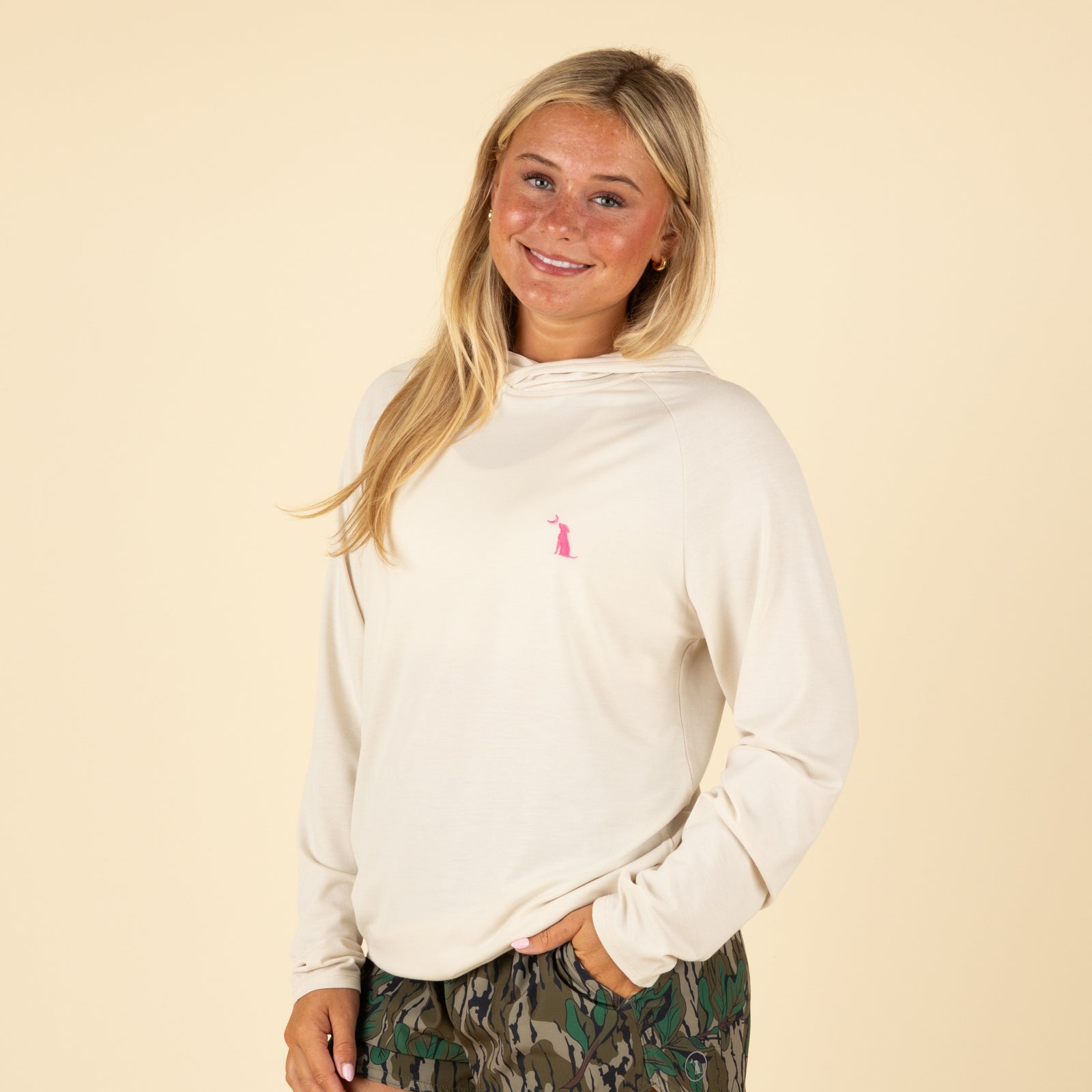 Woman wearing a cream hooded long sleeve shirt with pink dog logo on chest and green camouflage shorts, smiling with one hand in pocket.