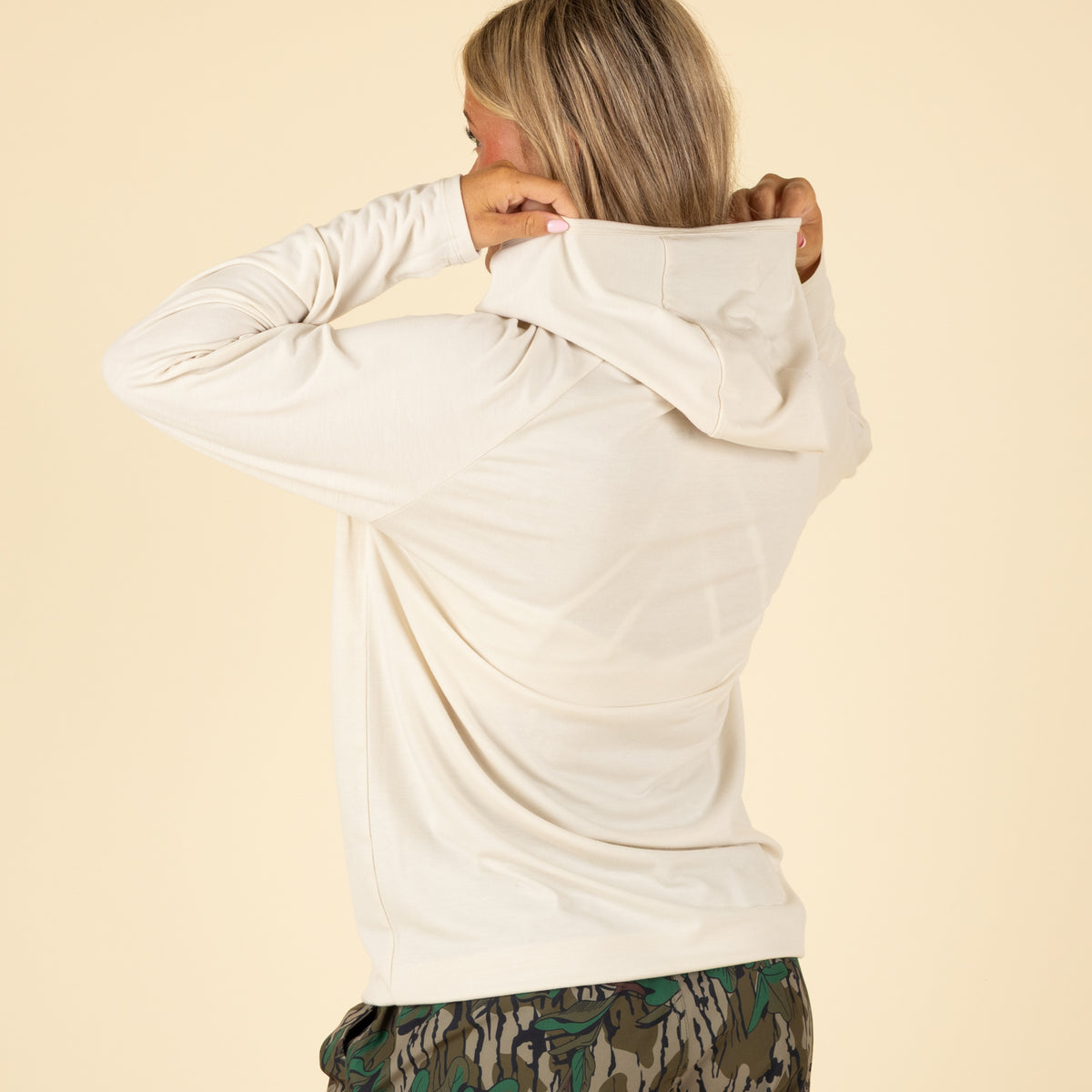 Back view of woman adjusting hood on cream long sleeve shirt, wearing green camouflage shorts.