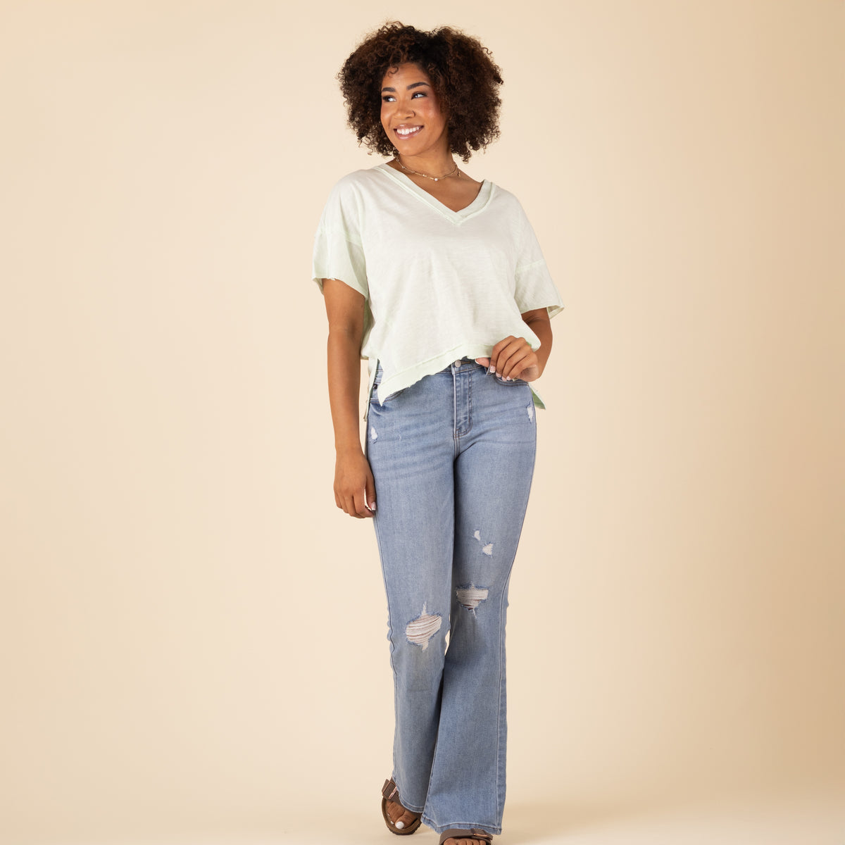 Model smiling in light green V-neck top and light-wash distressed flare jeans with ripped knees and frayed holes. Outfit styled with sandals.