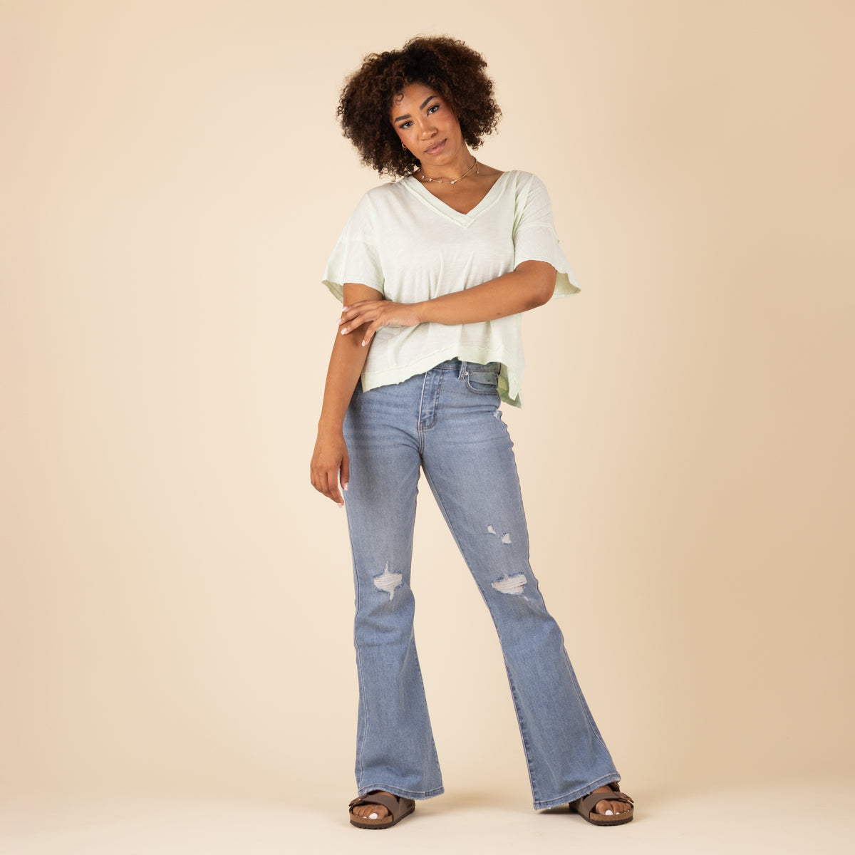 Model wearing light green V-neck top and light-wash flare jeans with knee distressing, standing casually with arms crossed and relaxed expression. 