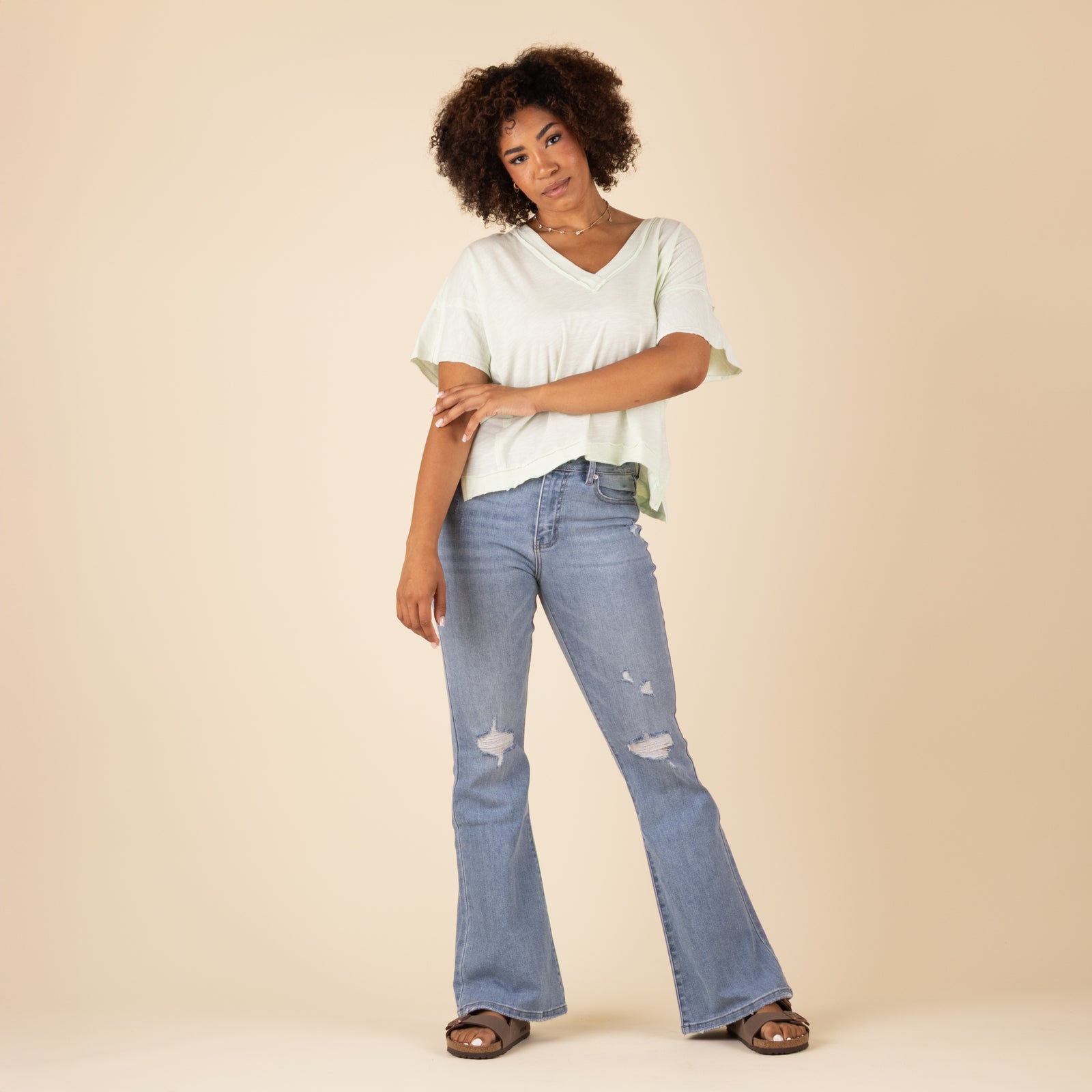 Model wearing light green V-neck top and light-wash flare jeans with knee distressing, standing casually with arms crossed and relaxed expression. 