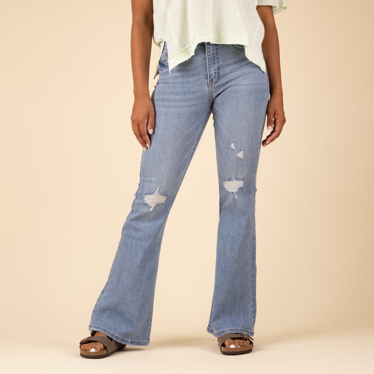 Woman wearing light-wash distressed flare jeans with front thigh rips and a light green V-neck top. Jeans have a high-rise fit and frayed holes. 