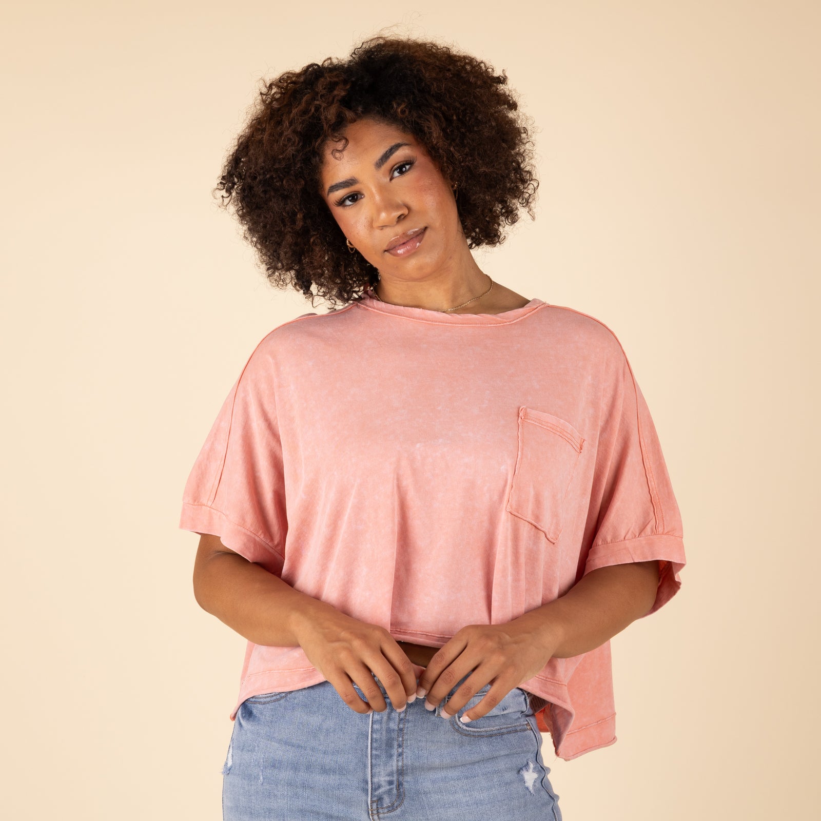 Very J Boxy Pocket Short Sleeve Crop Top, sizes S-L, color peach. Woman facing forward in a peach cropped t-shirt with a relaxed fit and front pocket, paired with jeans.