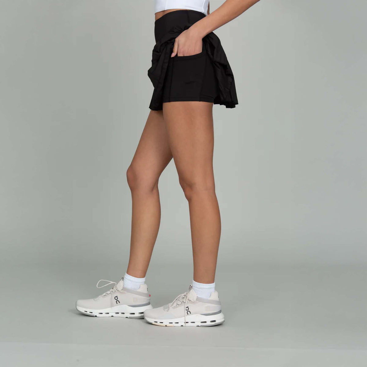 Side View Gold Hinge Pleated High-Waisted Tennis Skort in Black with hand in pocket of built in short liner