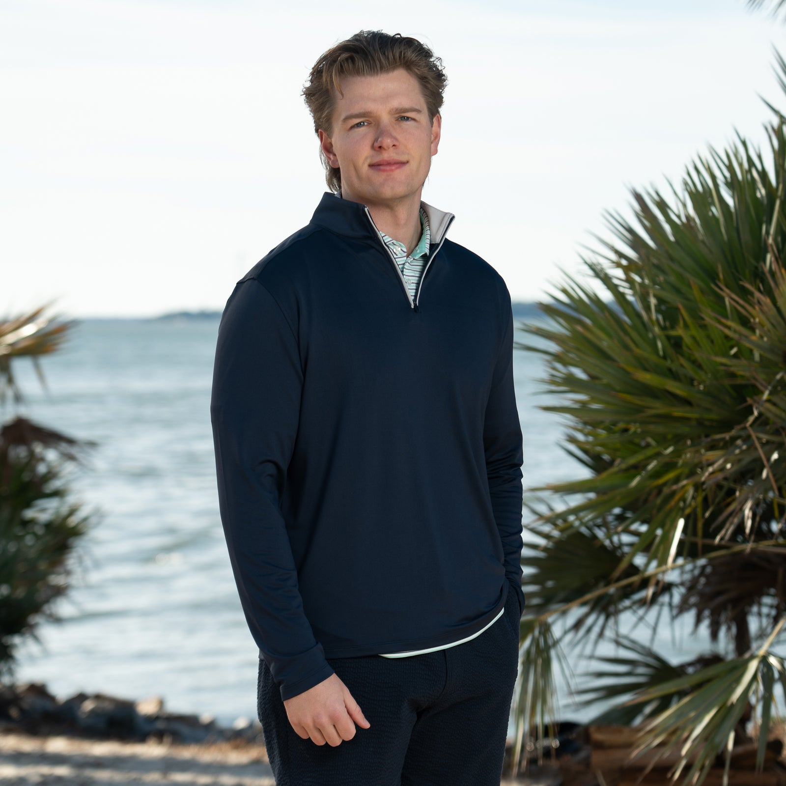 A man wearing a COAST Men's Mariner 1/4 Zip Pullover on a waterline with palm trees nearby, color Navy, Sizes S-XXL.  Designed with a sleek, athletic fit, it features a high collar and quarter-length zipper for adjustable coverage and easy layering. The soft, breathable fabric keeps you warm without weighing you down.