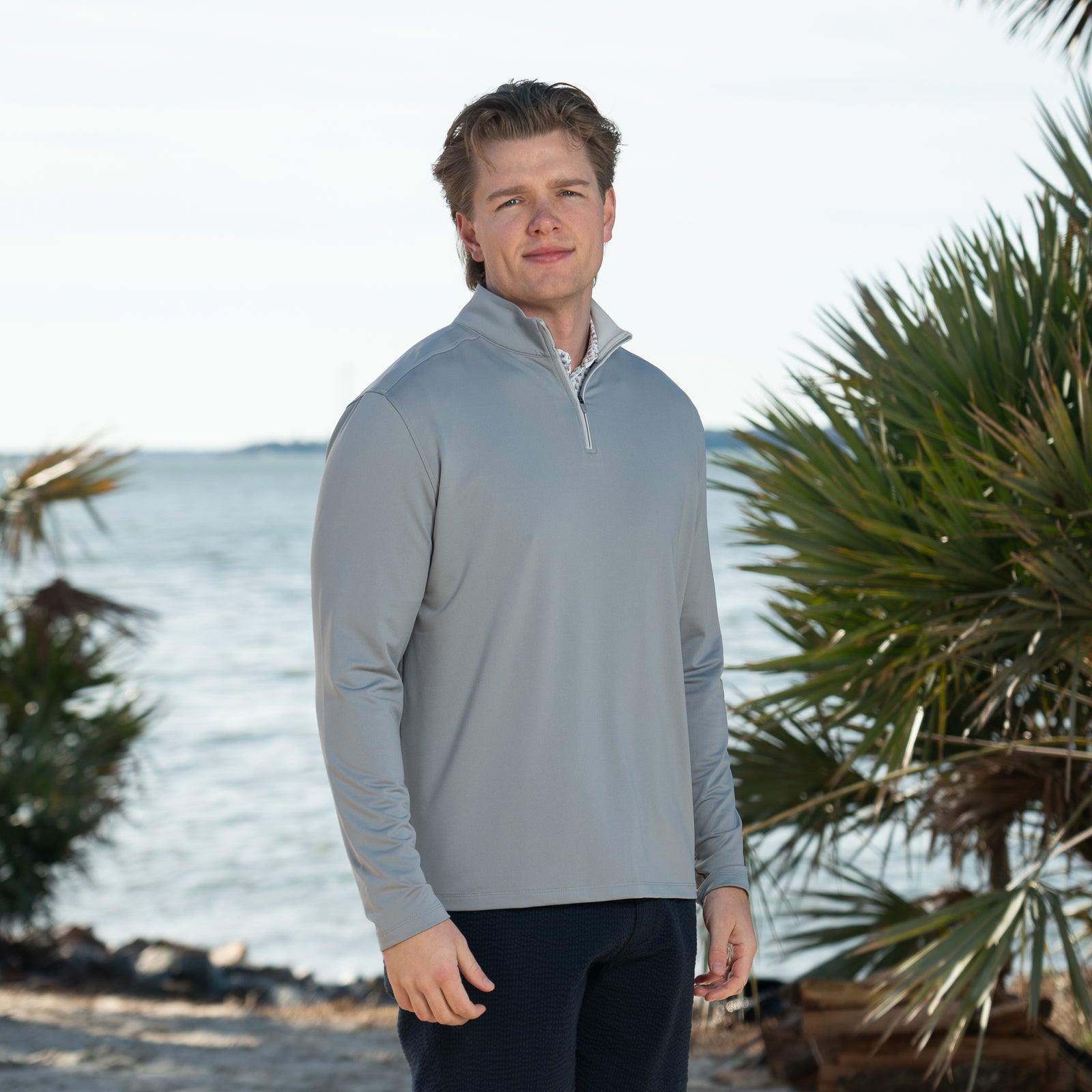 A man wearing a COAST Men's Mariner 1/4 Zip Pullover on a waterline with palm trees nearby, color Shark Fin Gray, Sizes S-XXL.  Designed with a sleek, athletic fit, it features a high collar and quarter-length zipper for adjustable coverage and easy layering. The soft, breathable fabric keeps you warm without weighing you down.