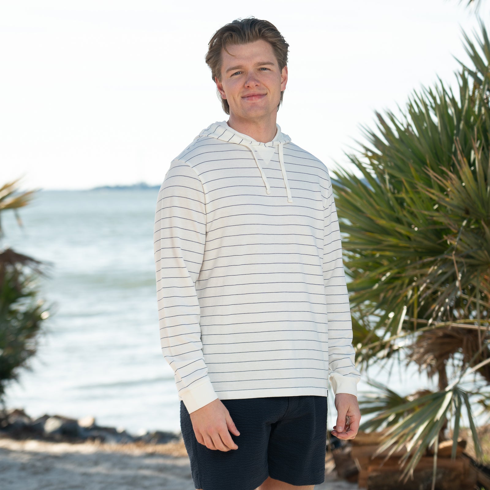 Coast Baja Men's Pullover in Ivory, sizes S-XXL, drawstring hood.
A man stands outdoors near the shoreline wearing a lightweight, long-sleeve ivory hoodie with thin navy horizontal stripes and a drawstring hood. He is also wearing dark navy textured shorts. Palm trees and calm water are visible in the background, suggesting a beachside setting. The man is facing the camera and smiling slightly in natural daylight.