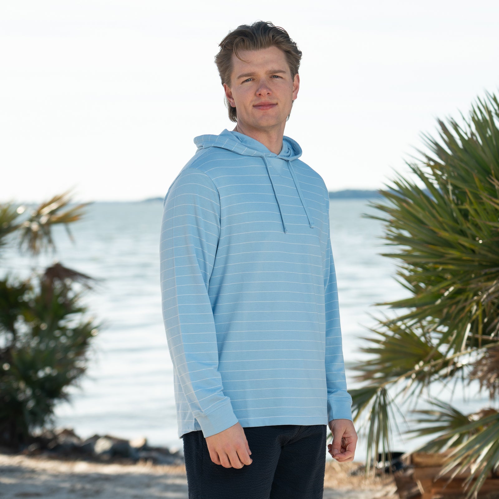 Coast Baja Men's Long Sleeve Pullover, color maya blue, sizes S-XXL, drawstring hoodie.
A man stands outdoors near the shoreline wearing a lightweight, long-sleeve maya blue hoodie with thin ivory horizontal stripes and a drawstring hood. He is also wearing dark navy textured shorts. Palm trees and calm water are visible in the background, suggesting a beachside setting. The man is facing the camera and smiling slightly in natural daylight.
