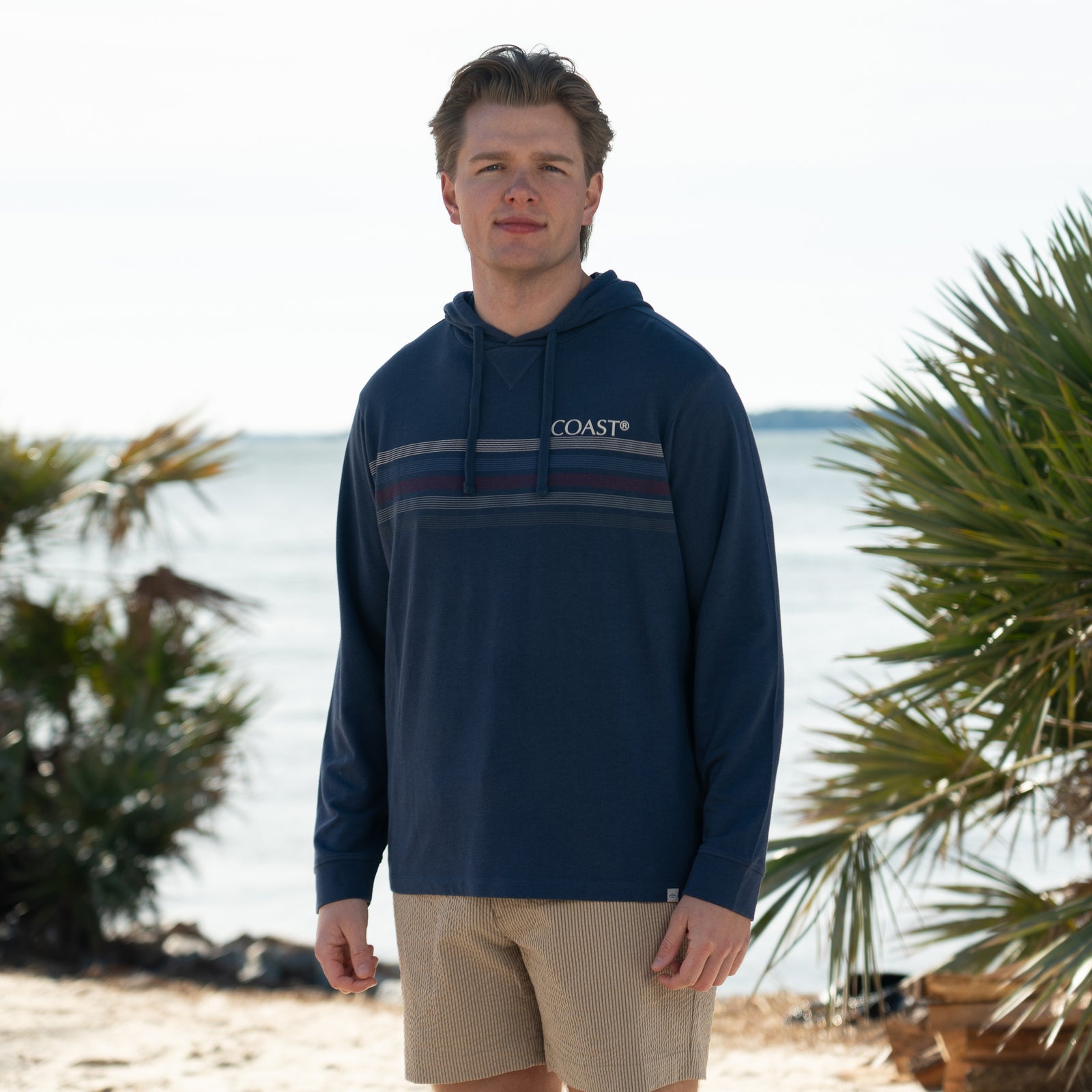 Coast Baja Men's Long Sleeve pullover in Maya blue, sizes S-XXL, drawstring hoodie.
A man stands outdoors by the shoreline wearing a lightweight, long-sleeve slate blue hoodie with thin ivory, dark red, and blue horizontal stripes across the chest, with the Coast logo on the left chest, and a drawstring hood. He is also wearing khaki textured shorts. Palm trees and calm water are visible in the background, suggesting a beachside setting. The man is facing the camera and smiling slightly in natural daylight.