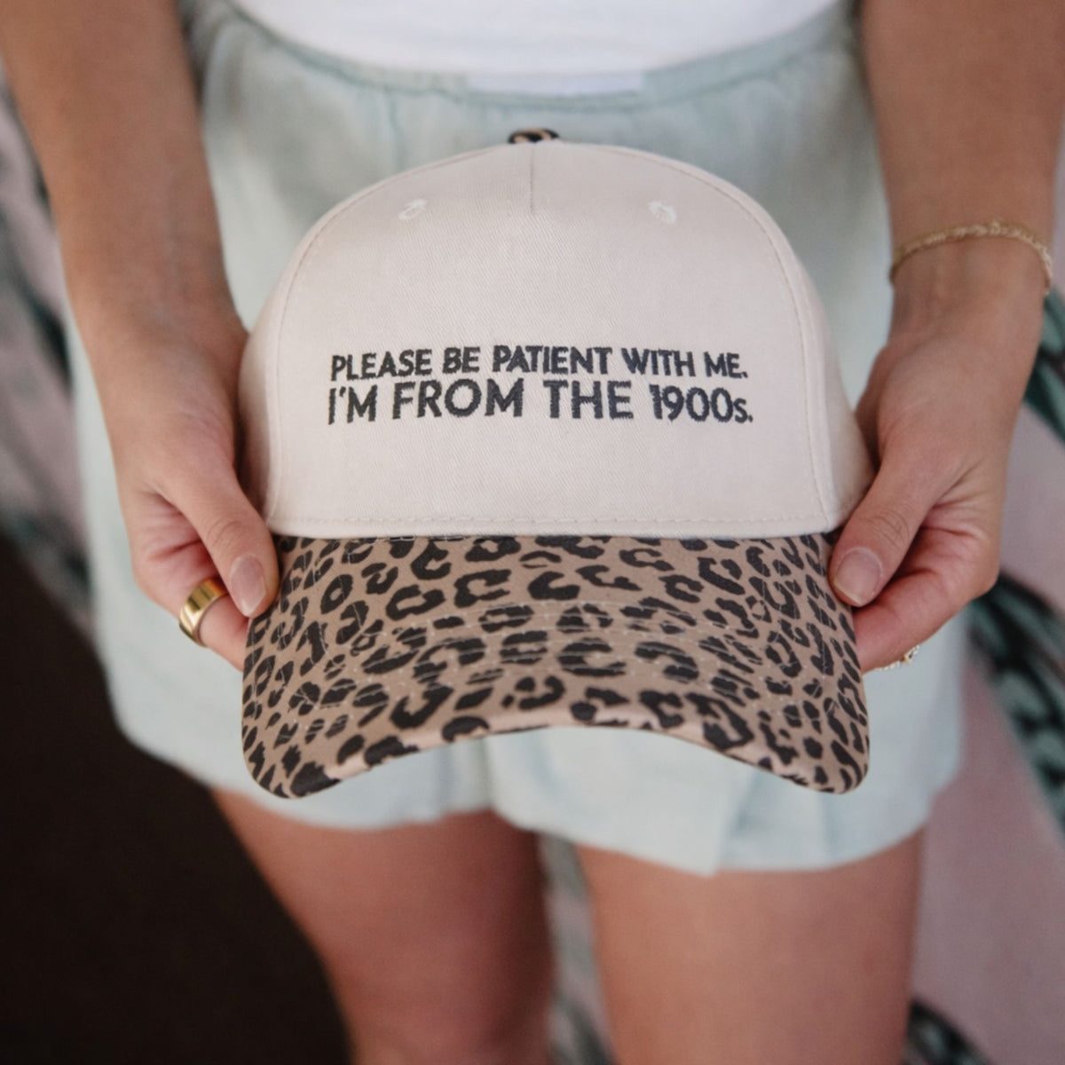 Tan trucker hat with black embroidered text and leopard print brim, held in hands showing front detail.
