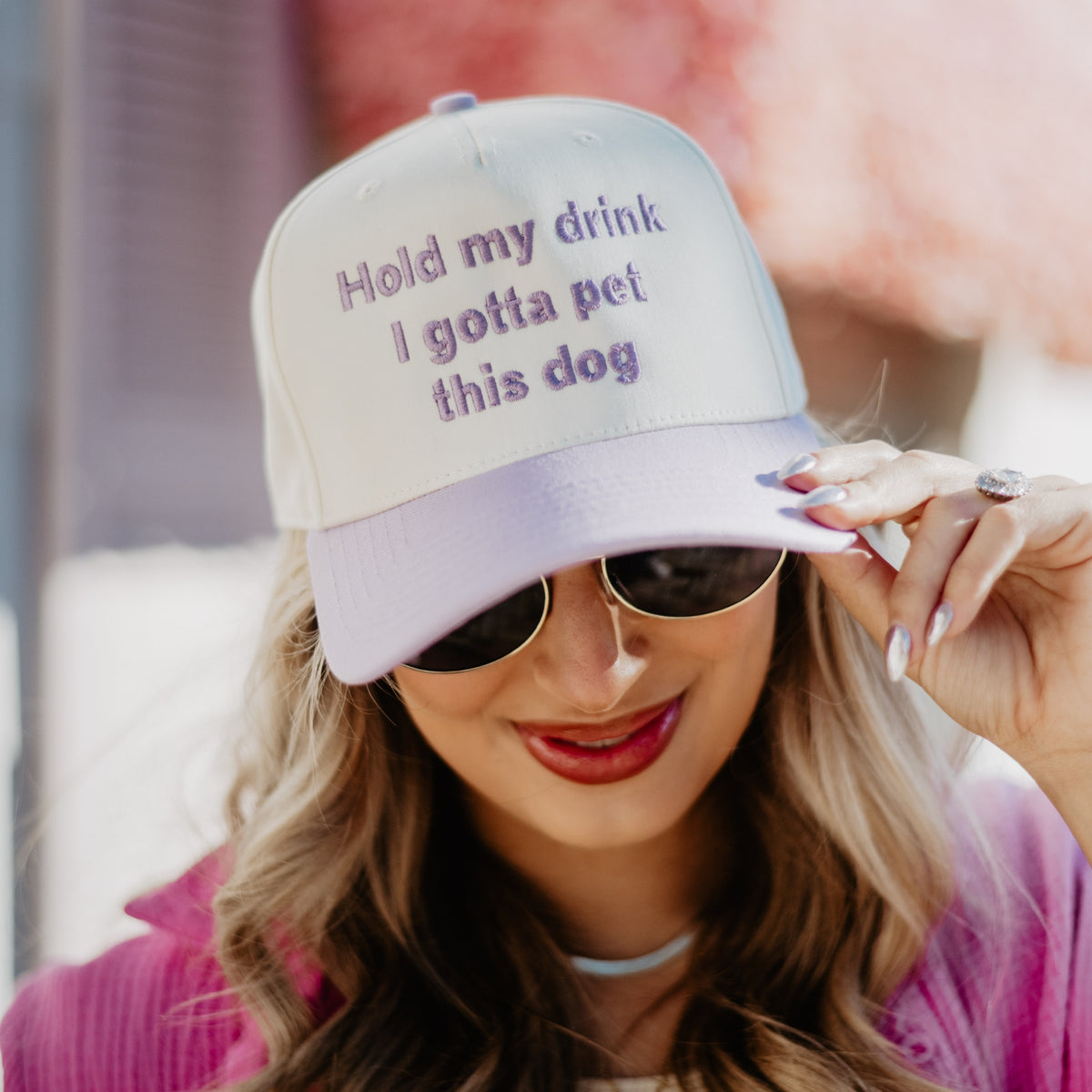 Tan and lavender trucker hat with “Hold my drink I gotta pet this dog” embroidered text, worn in lifestyle close-up view.