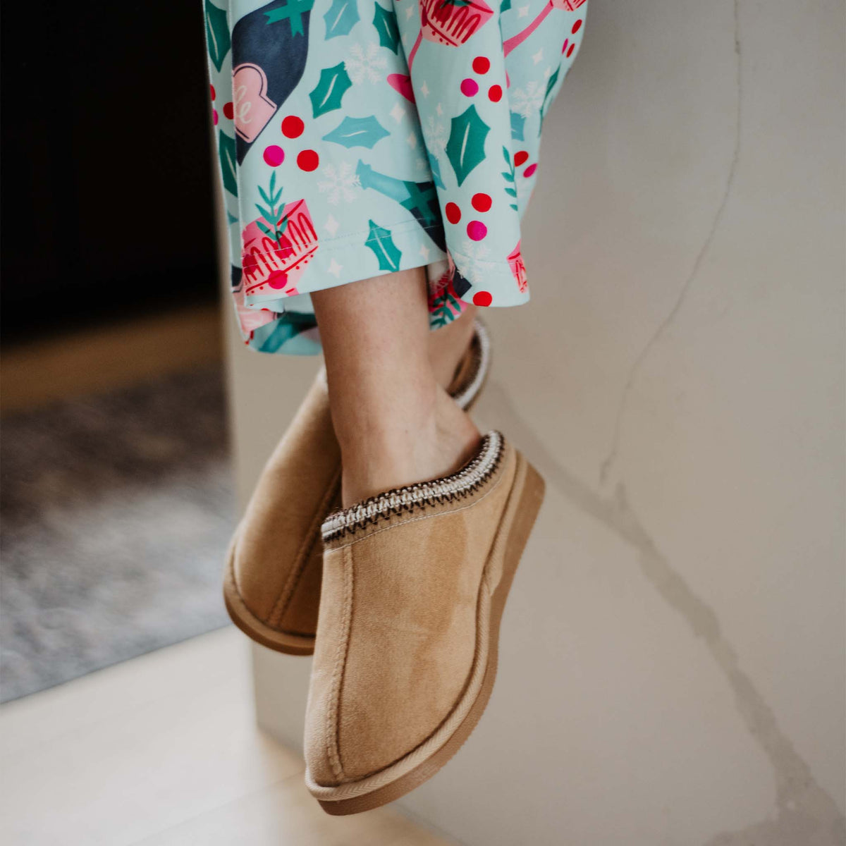 Lifestyle image showing tan slip-on slippers worn indoors with patterned pajama pants.