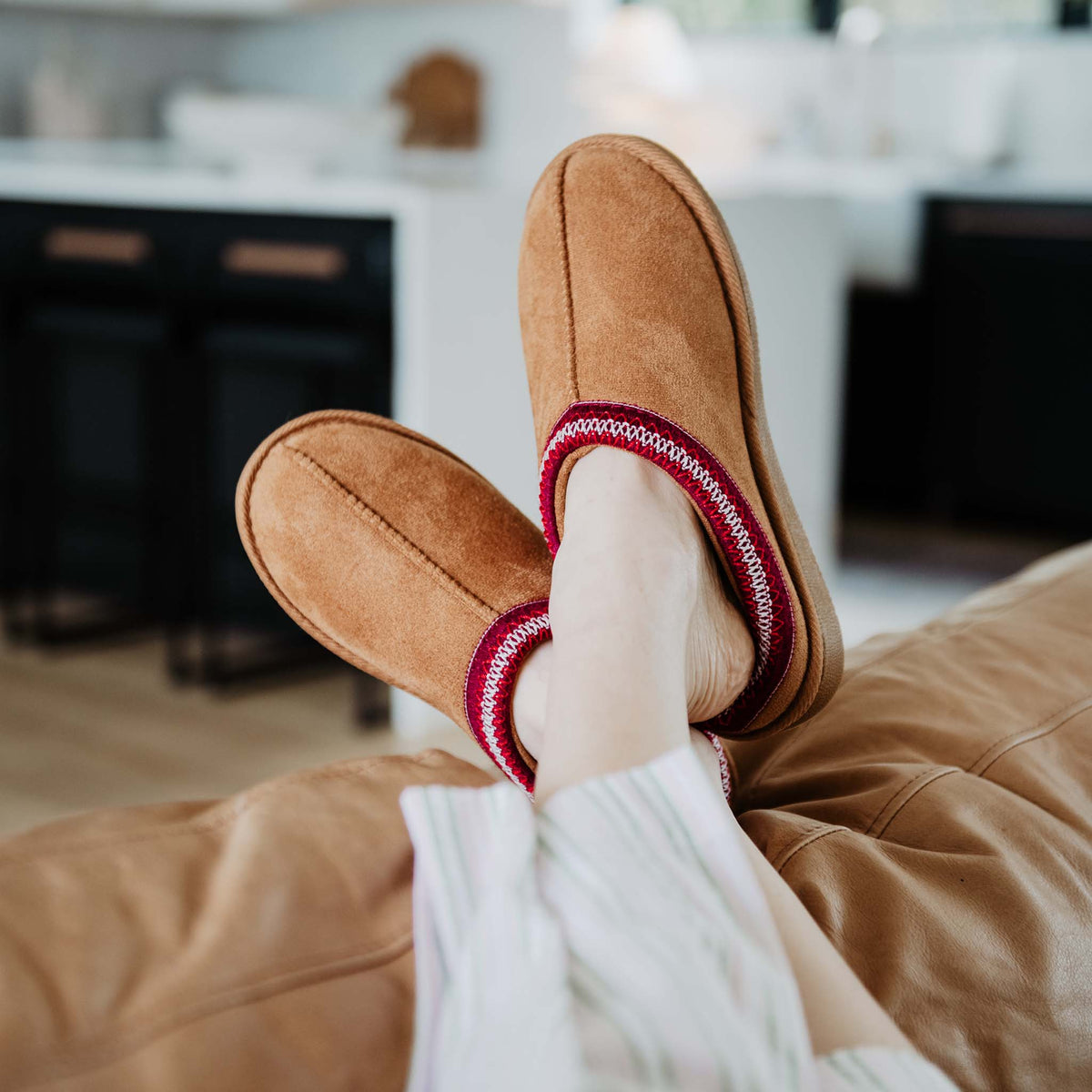Lifestyle image of chestnut slip-on slippers with red stitched trim, worn while lounging indoors.