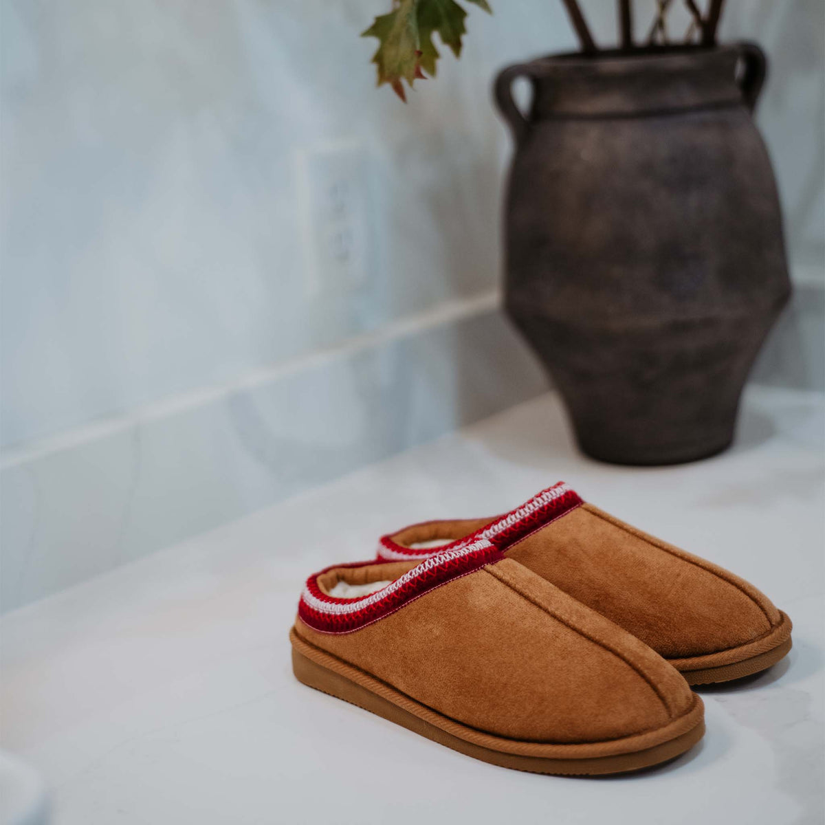 Chestnut slip-on slippers with red stitched trim displayed on a countertop near a decorative vase.