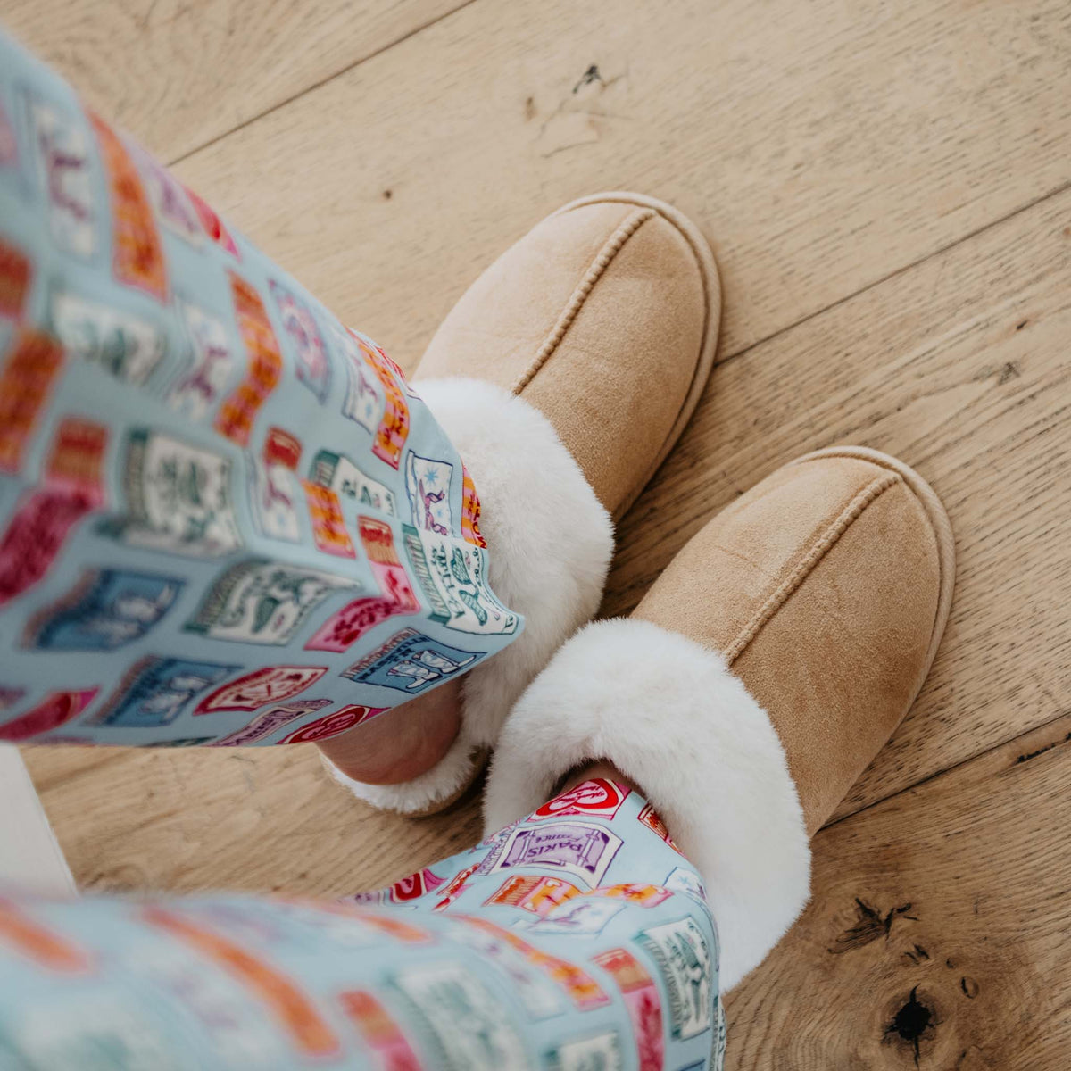 Lifestyle image of tan slippers with white faux fur trim worn indoors with patterned pajama pants on wood flooring.