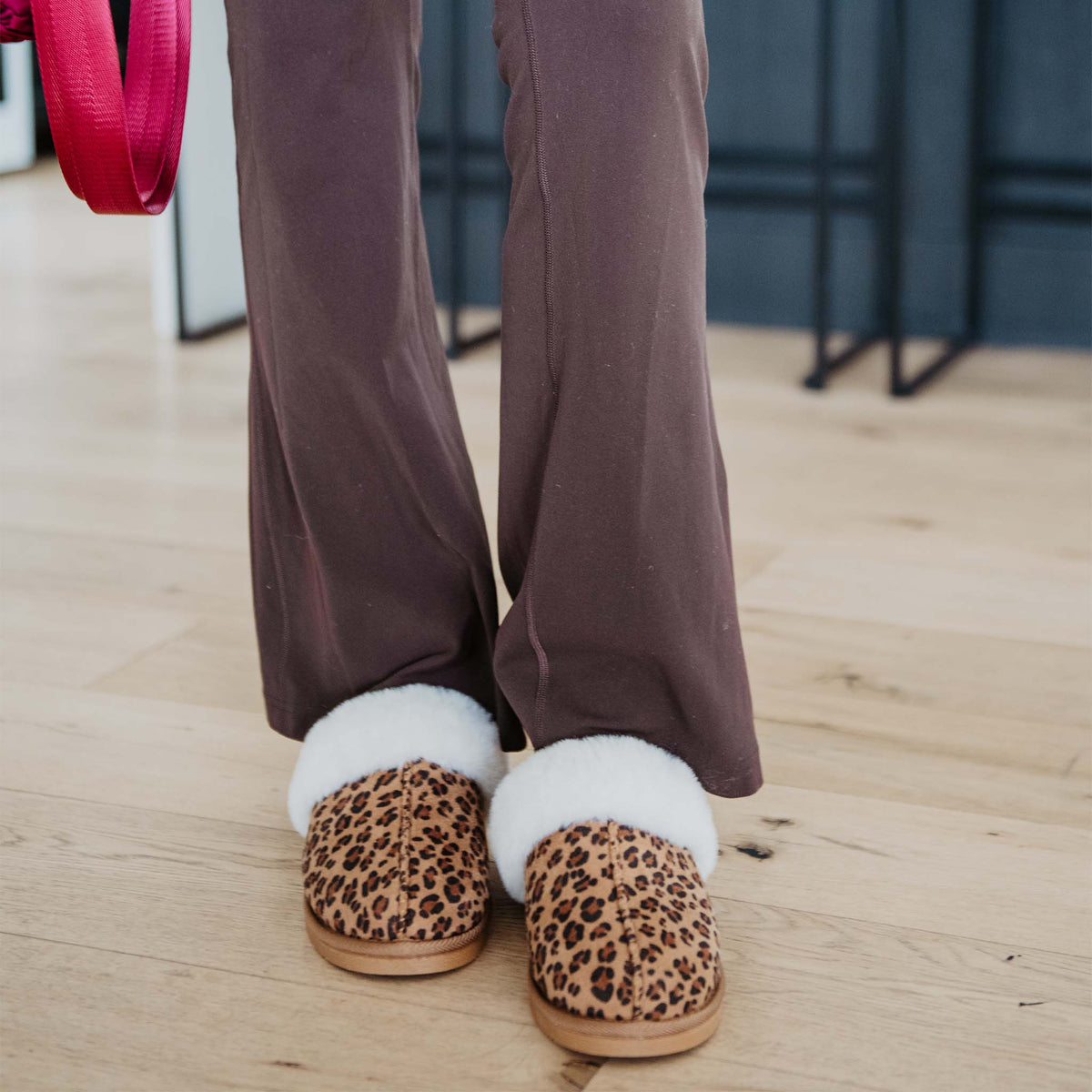 Lifestyle image of leopard print slippers with white faux fur trim worn indoors with brown pants on wood flooring.