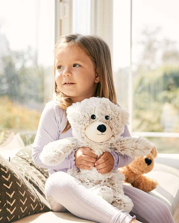 a little girl sitting by a window holding a Warmies bear stuffed animal