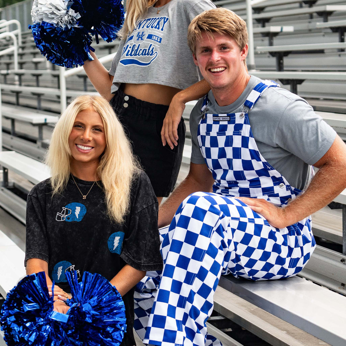 Man in blue and white checkered overalls sits in stadium stands with women wearing Kentucky gear and holding blue pom-poms, all smiling on game day.