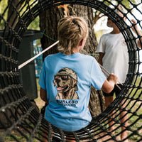 Burlebo Outdoors youth light blue short sleeve t-shirt with dog graphic wearing sunglasses and camo hat on back, shown on child outdoors in a rope playground.