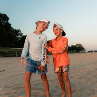 Two boys wearing long sleeve performance shirts and swim trunks laughing together on a sandy beach at sunset.