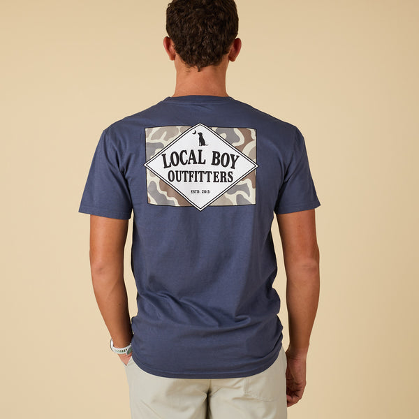 Man wearing a navy blue t-shirt with 'Local Boy Outfitters' logo on the back against a beige background