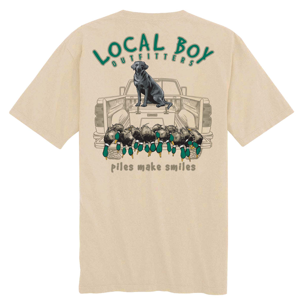 Back view of tan short sleeve t-shirt with Local Boy Outfitters logo, black lab on truck tailgate, and caption “Piles Make Smiles” below ducks.
