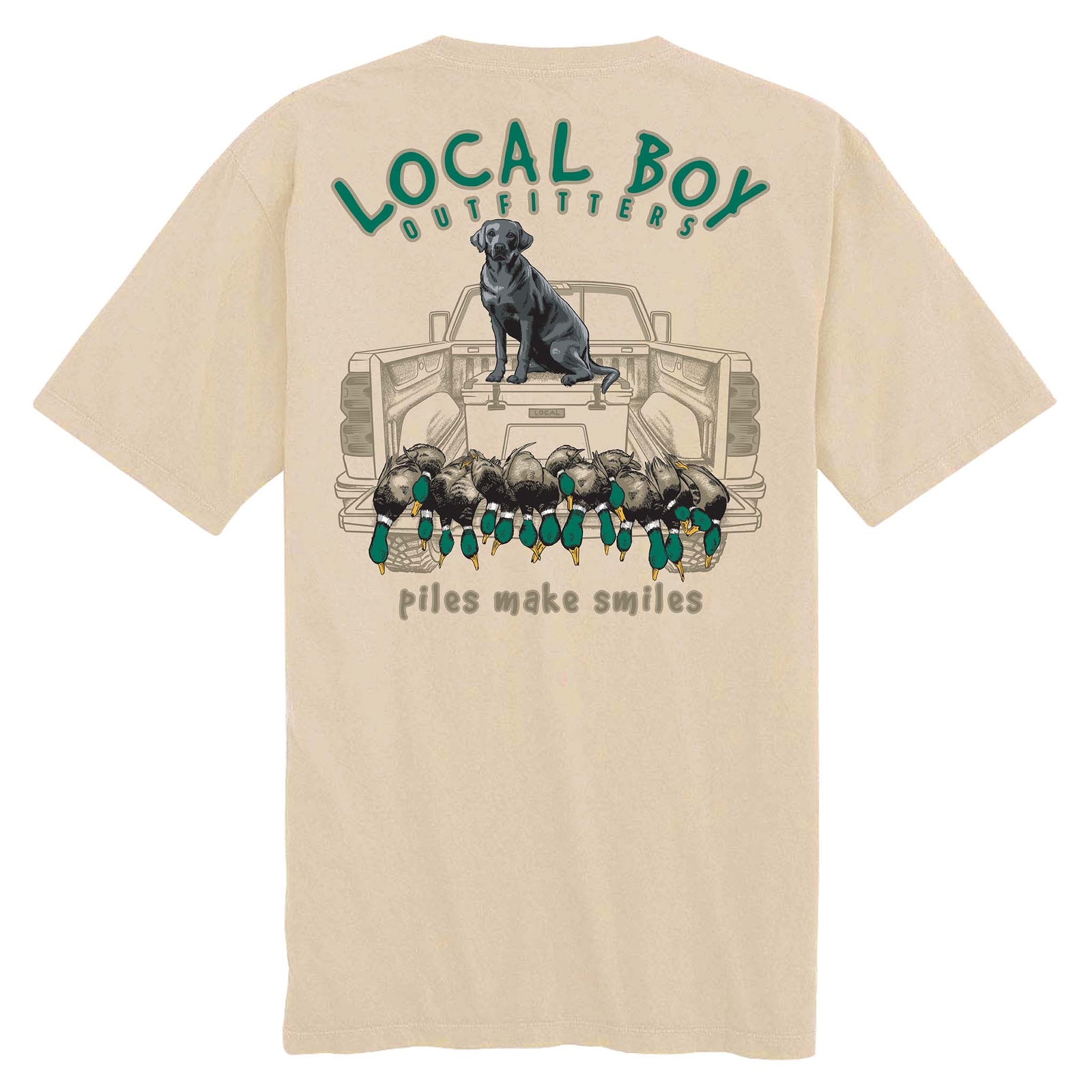 Back view of tan short sleeve t-shirt with Local Boy Outfitters logo, black lab on truck tailgate, and caption “Piles Make Smiles” below ducks.