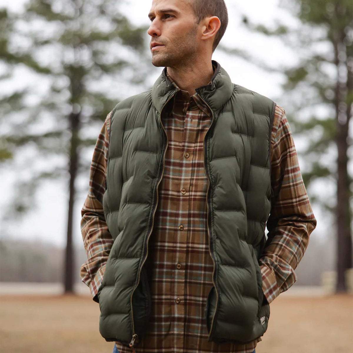 Man wearing a green puffer vest over a plaid shirt in a forest setting