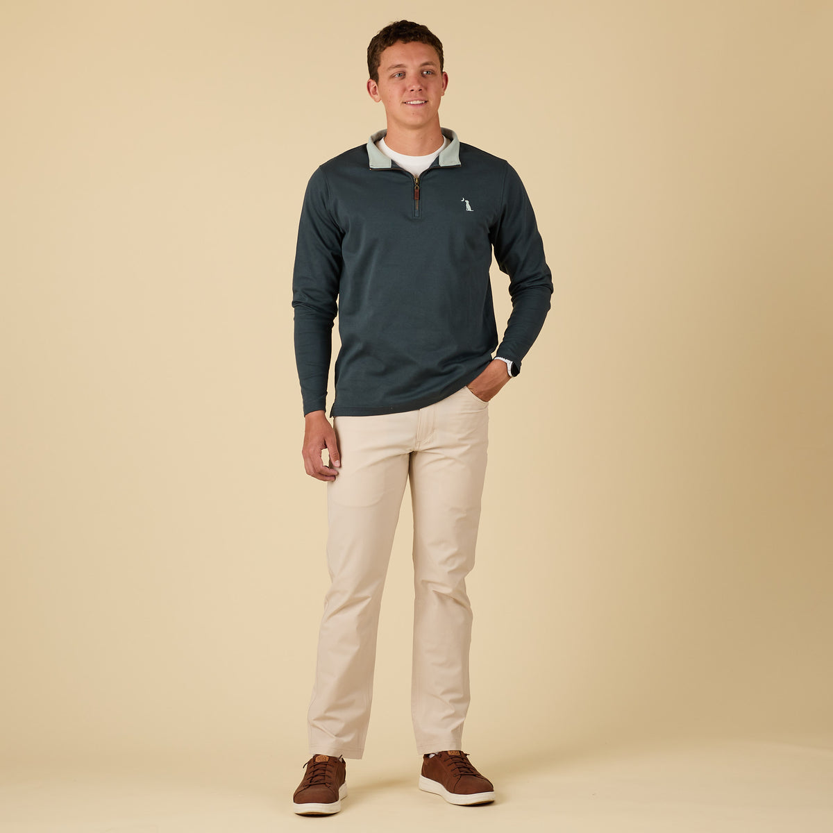 Man wearing a dark blue pullover with a logo, beige pants, and brown shoes on a beige background